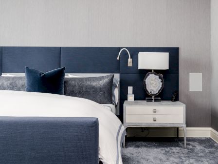 A modern bedroom with a navy upholstered headboard, white bedding, and a bedside table with a lamp and decorative item.