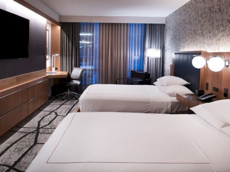 A modern hotel room with two beds, a TV, and a desk near the window, featuring stylish decor and ambient lighting.