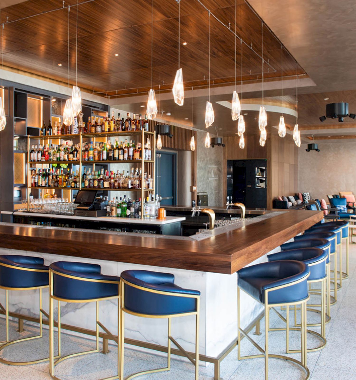 This is a modern bar with a long wooden counter, blue bar stools, and a selection of bottles. Elegant lighting hangs overhead.