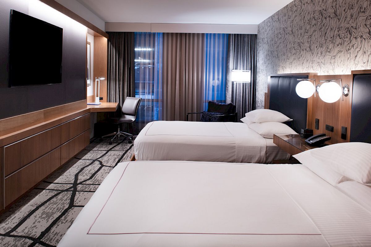 A modern hotel room with two beds, a desk, a wall-mounted TV, and stylish decor.