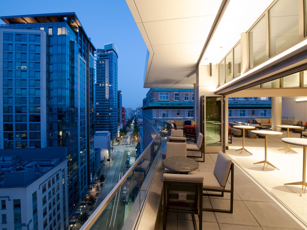 A modern rooftop patio with seating overlooks a city's high-rise buildings and illuminated street at dusk, showcasing urban nightlife.