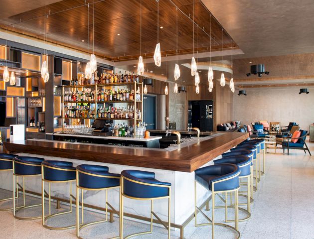 A modern bar features a well-stocked counter, stylish hanging lights, and comfortable seating in a spacious, contemporary setting.