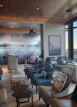 The image shows a modern lounge with seating, a mural on the wall, and large windows offering a view of water and a Ferris wheel outside.