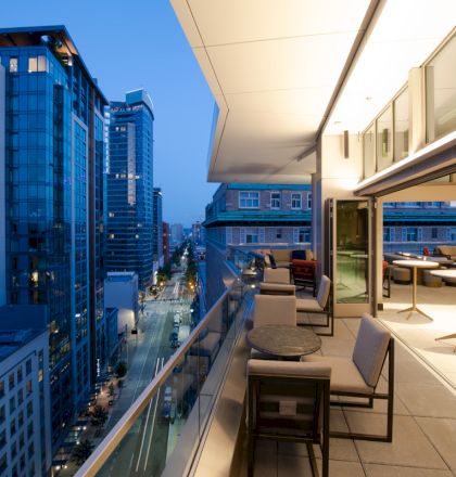 A modern rooftop patio with tables and chairs overlooks a city street lined with tall buildings at dusk.