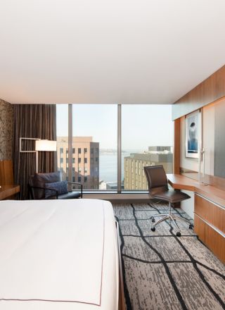 A modern hotel room with a large bed, desk, chair, and TV, overlooking a cityscape through wide windows.