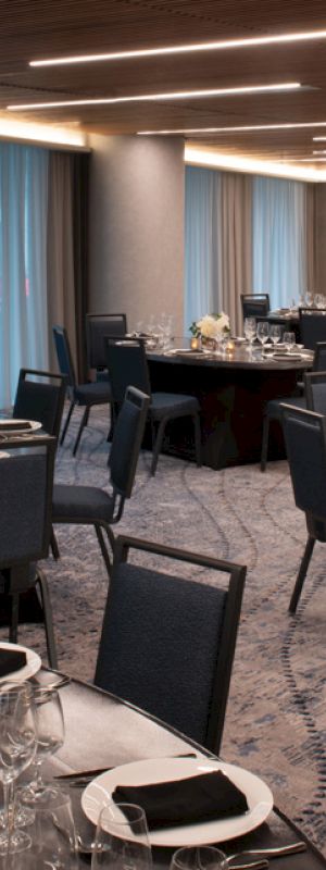 The image shows a dimly lit banquet hall with round tables set for dining, featuring black chairs and white floral centerpieces.
