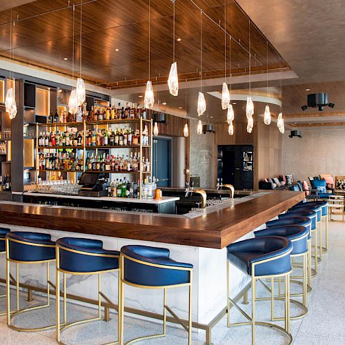 A modern bar with a wooden counter, blue chairs, hanging lights, and well-stocked shelves is featured in a spacious lounge setting.