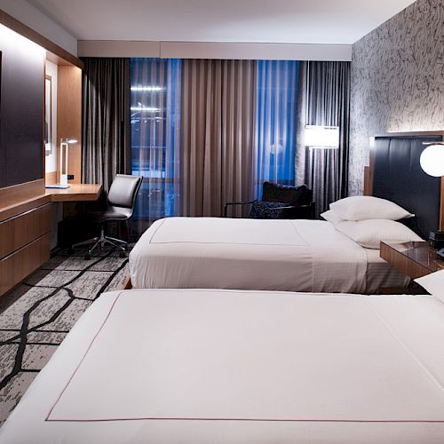 The image shows a modern hotel room with two beds, a wall-mounted TV, a desk and chair, and stylish decor.