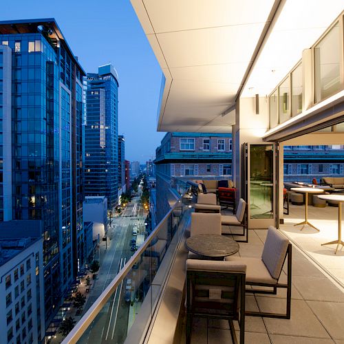 A modern rooftop terrace with seating overlooks city buildings at dusk, with street lights illuminating the scene.