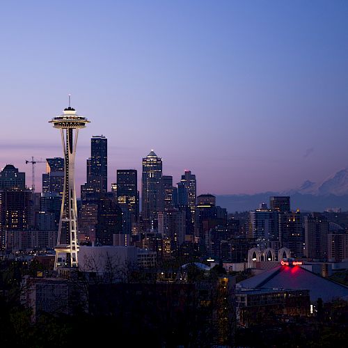The image shows the Seattle skyline at dusk, featuring the Space Needle and Mount Rainier in the background, with city lights illuminating.
