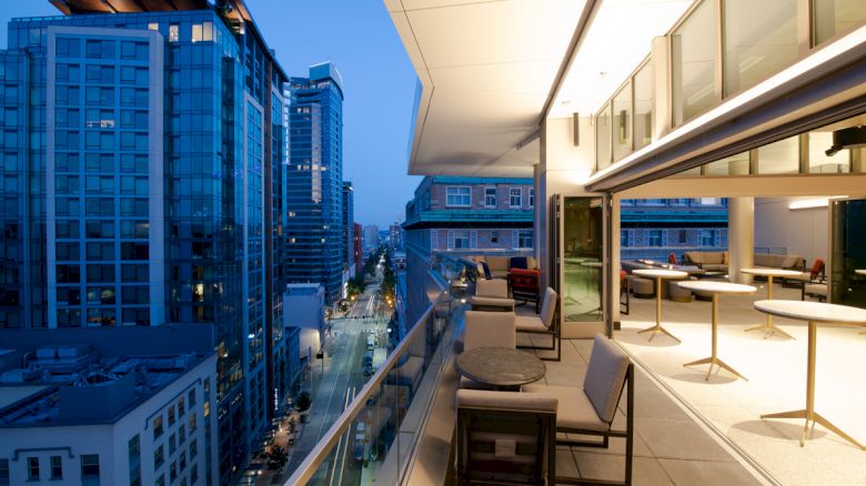 A modern cityscape view from a balcony at dusk, featuring tall buildings, a street below, and an outdoor seating area.