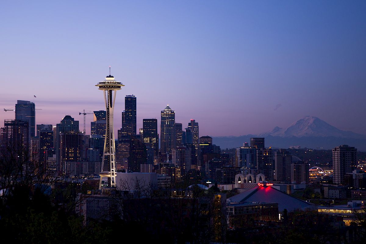 A nighttime cityscape with illuminated buildings and a backdrop of mountains, featuring a prominent tower with a disk-shaped top.