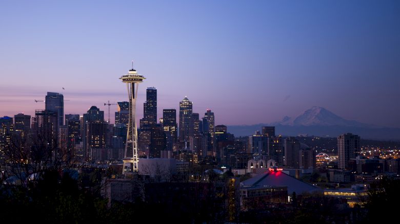 A nighttime cityscape with illuminated buildings and a backdrop of mountains, featuring a prominent tower with a disk-shaped top.