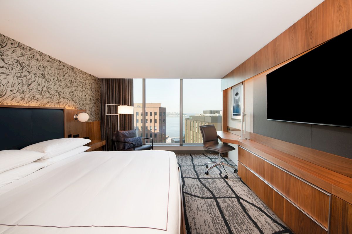 A modern hotel room with a large bed, wall-mounted TV, desk, chair, and city view through the window.