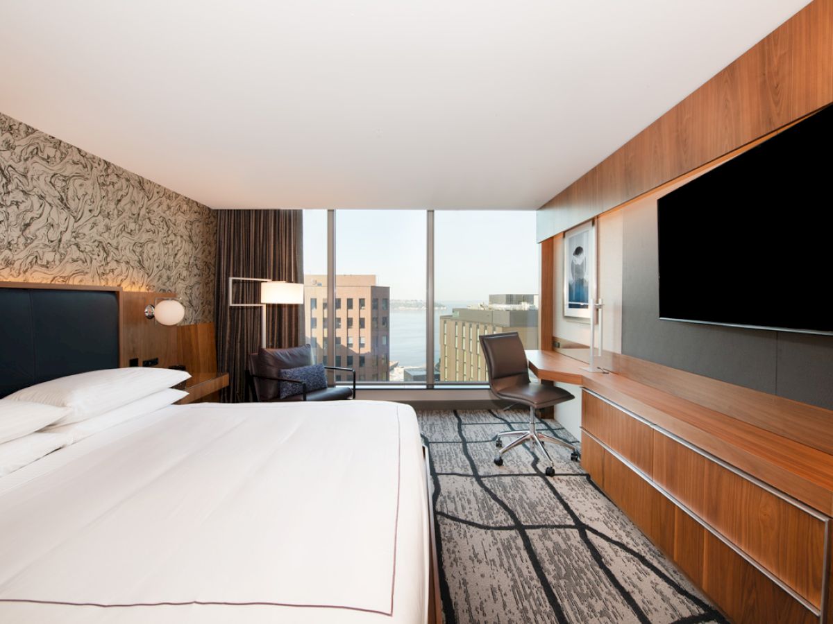 A modern hotel room with a large bed, wall-mounted TV, desk, chair, and city view through the window.