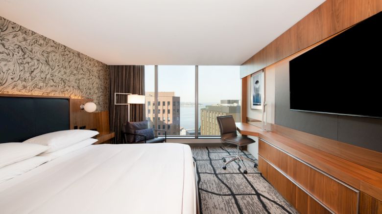 A modern hotel room with a large bed, wall-mounted TV, desk, chair, and city view through the window.