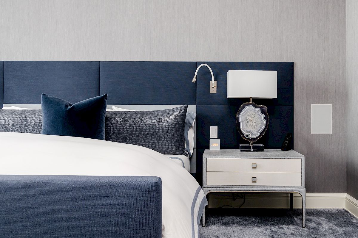 A modern bedroom with a blue upholstered bed, navy pillows, gray bedding, a white nightstand, and a lamp on a gray carpet.