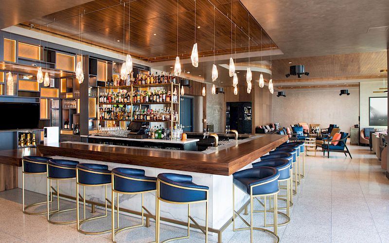 A modern bar with blue stools, a well-stocked shelf, hanging lights, and a spacious seating area with cozy chairs and tables.